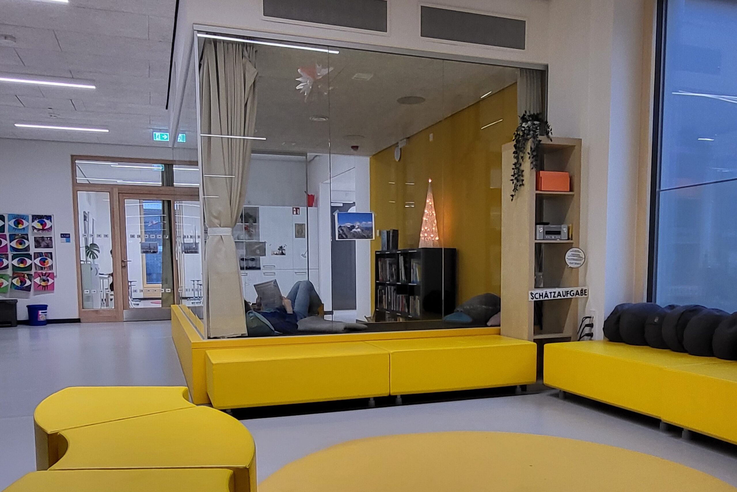 modern gestalteter Raum an Grundschule mit Sitzelementen, Glaskubus, in dem jemand gemütlich liest
